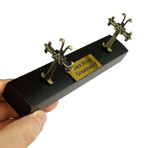 Op de afbeelding: Een decoratief display met twee bronzen zwaardreplica's, elk met een sierlijk kruisvormig handvat. Ze zijn bevestigd op een zwarte houten basis, met een goudkleurig plaatje met de tekst "Dark Moon Greatsword".