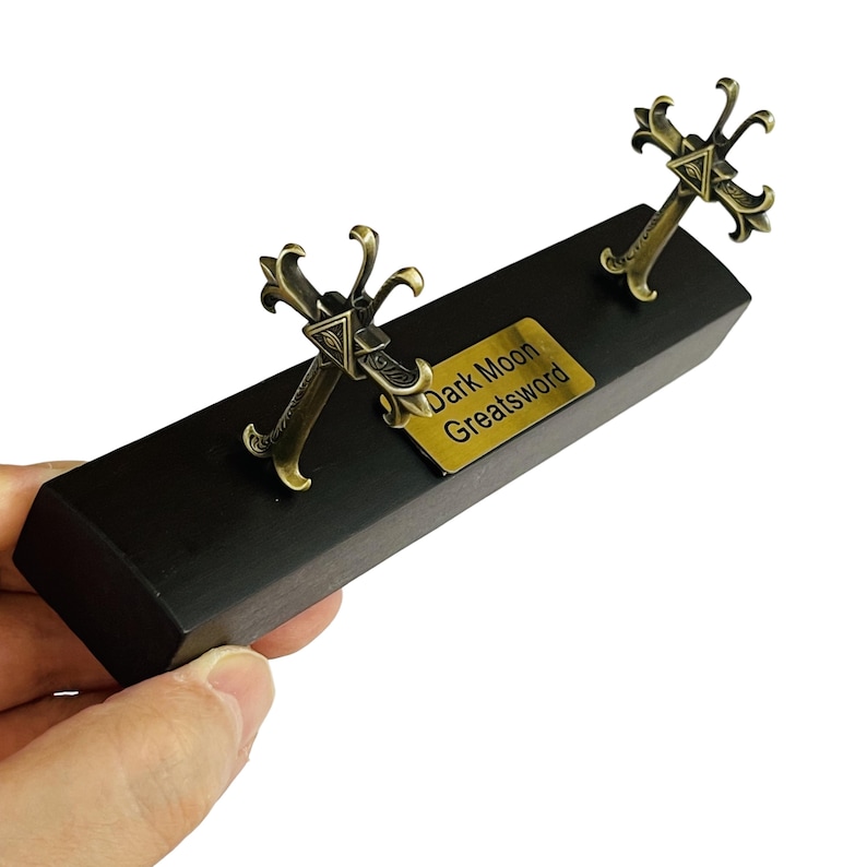 Op de afbeelding: Een decoratief display met twee bronzen zwaardreplica's, elk met een sierlijk kruisvormig handvat. Ze zijn bevestigd op een zwarte houten basis, met een goudkleurig plaatje met de tekst "Dark Moon Greatsword".