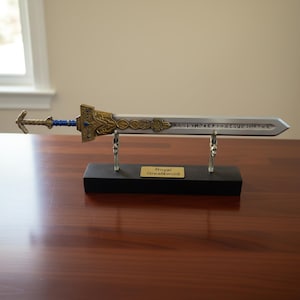 Op de afbeelding: Een decoratieve replica van het Koninklijke Grootzwaard. Het zwaard heeft een zilveren kling met gegraveerde inscripties, een gouden en blauwe pareerstang en wordt tentoongesteld op een zwarte standaard met een gouden naamplaat. Het zwaard is ongeveer 76 cm lang.