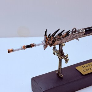 May include: A detailed miniature sword with a dragon-themed hilt and blade. The sword features a bronze and black color scheme, with intricate detailing on the handle and guard. The blade is black and the handle is white. The sword is mounted on a black base.