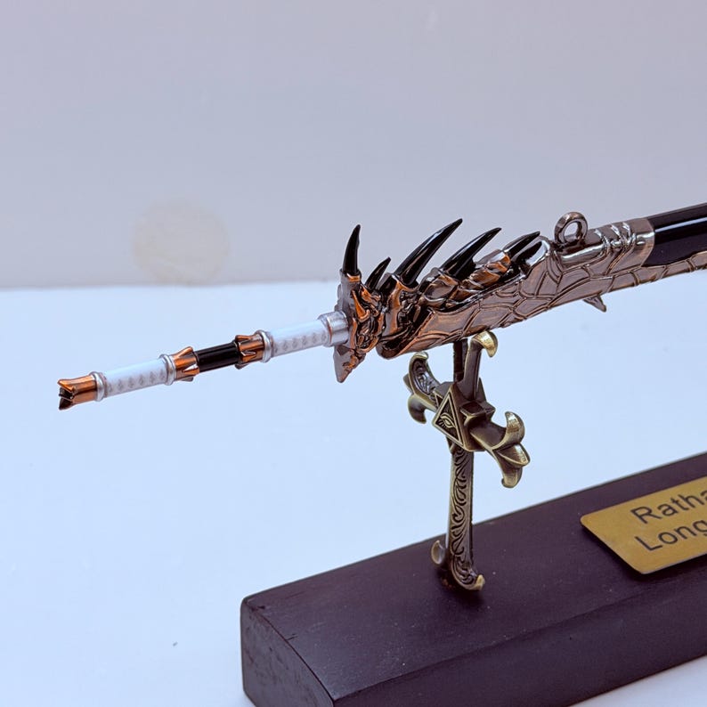 May include: A detailed miniature sword with a dragon-themed hilt and blade. The sword features a bronze and black color scheme, with intricate detailing on the handle and guard. The blade is black and the handle is white. The sword is mounted on a black base.