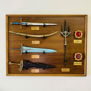 Op de afbeelding: Een houten display met verschillende fantasiewapens, waaronder zwaarden en een speer. De display bevat een zwaard met het label "Carian Knight's Sword", een gebogen zwaard met het label "Malenia's Freehand Sword" en een zwaard met blauw lemmet met het label "Holy Moonlight Sword".