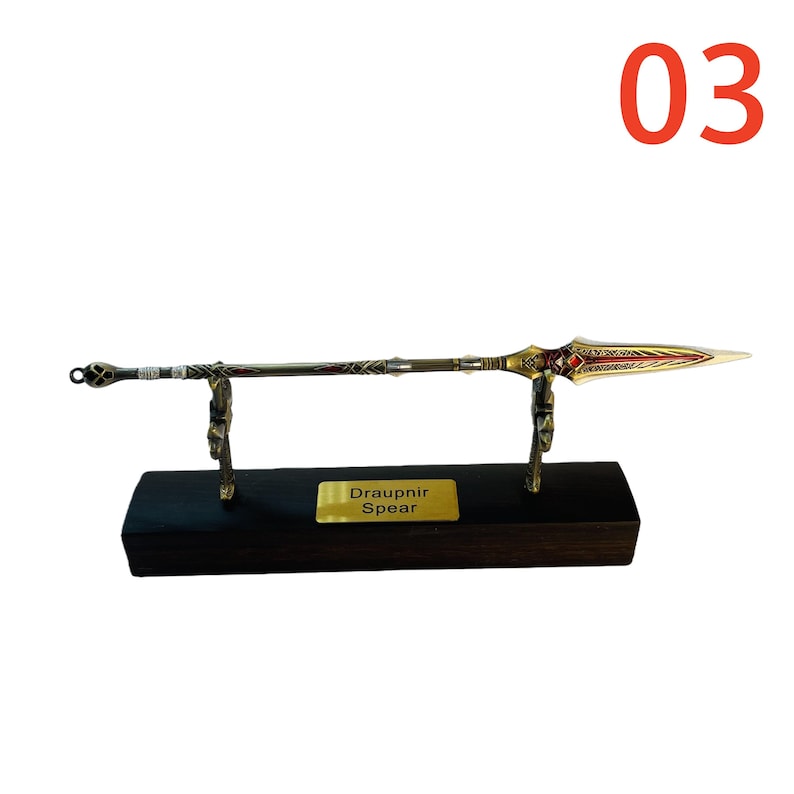 May include: A decorative spear, the Draupnir Spear, is displayed on a black wooden base. The spear features a gold-colored blade with red accents and a detailed handle. The number "03" is in the upper right corner.