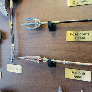 May include: A display of fantasy weapons, including a trident with blue tines and a spear with red and silver accents. The sign reads "Poseidon's Trident" and "Draupnir Spear". The weapons are mounted on a brown wall.