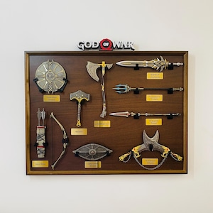 May include: A wooden display case featuring replicas of weapons from the "God of War" video game. Includes a Talon Bow, Guardian Shield, Mjolnir, Leviathan Axe, Blade of Olympus, Poseidon's Trident, Draupnir Spear, Dauntless Shield, and Blades of Chaos.