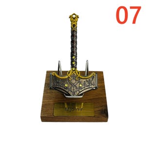 May include: A decorative Mjolnir hammer replica, featuring a detailed gold and silver design with embedded yellow gems. The hammer rests on a wooden base with a gold nameplate that reads "Mjolnir". The number "07" is in the upper right corner.
