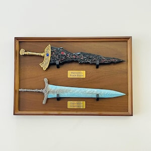 Op de afbeelding: Een houten display met twee decoratieve zwaarden. Het bovenste zwaard, "Maliketh's Black Blade", is zwart met gouden accenten. Het onderste zwaard, "Dark Moon Greatsword", is lichtblauw en zilver.