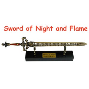 Op de afbeelding: Een decoratief zwaard met een bronzen afwerking, tentoongesteld op een zwarte standaard. Het zwaard heeft ingewikkelde details en een rode handgreep. De tekst "Sword of Night and Flame" is zichtbaar boven het zwaard.