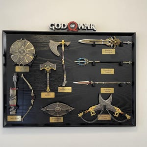 May include: A black framed display featuring replicas of weapons from the God of War video game. Includes the Blade of Olympus, Leviathan Axe, Mjolnir, and Talon Bow. The God of War logo is at the top.