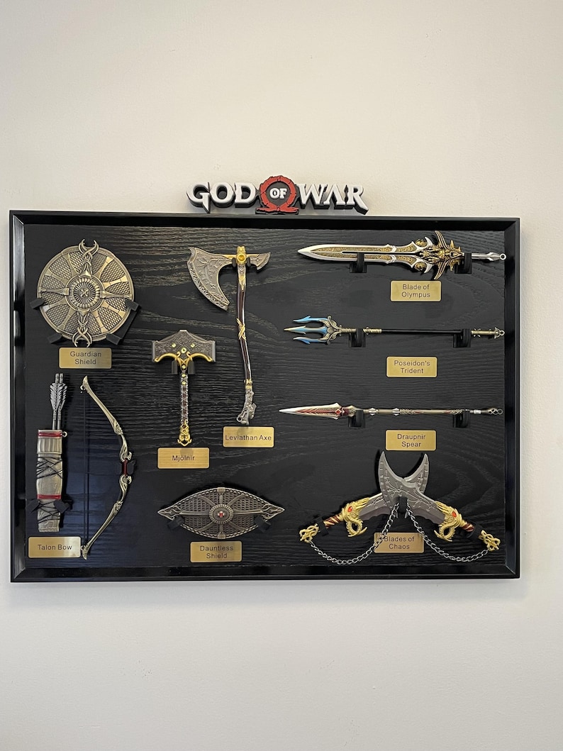 May include: A black framed display featuring replicas of weapons from the God of War video game. Includes the Blade of Olympus, Leviathan Axe, Mjolnir, and Talon Bow. The God of War logo is at the top.