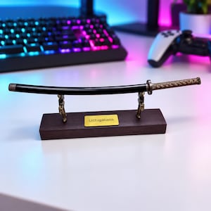 Op de afbeelding: Een miniatuur Uchigatana zwaardreplica met een zwarte schede en een getextureerd bronskleurig handvat. Het zwaard rust op een donkere houten standaard met twee metalen steunen en een goudkleurig plaatje met de tekst Uchigatana.