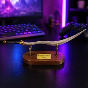 May include: A curved metal sword replica with a black wrapped handle and gold-colored accents, resting on a wooden display stand. The stand features a small rectangular gold plaque with the engraved text Bloodhound's Fang centered on the front.