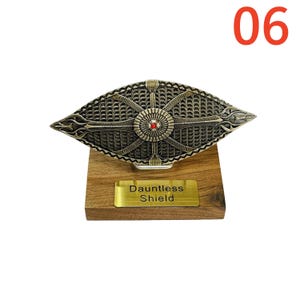 May include: A bronze-colored Dauntless Shield, featuring intricate geometric patterns and a central red gem, is mounted on a wooden base. A gold plaque on the base reads "Dauntless Shield". The number "06" is in the upper right corner.