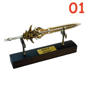 May include: A decorative sword with intricate gold and silver detailing, displayed on a wooden base. The sword is labeled "Blade of Olympus" on a small gold plaque. The number "01" is in the upper right corner.