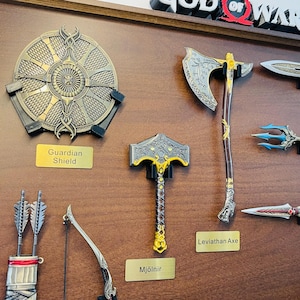May include: A collection of fantasy-themed weapons, including a shield, axe, and hammer, displayed on a wooden surface. The shield is round with intricate detailing. The axe and hammer have ornate designs. The text labels read "Guardian Shield", "Leviathan Axe", and "Mj&ouml;lner".