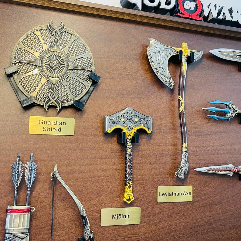 May include: A collection of fantasy-themed weapons, including a shield, axe, and hammer, displayed on a wooden surface. The shield is round with intricate detailing. The axe and hammer have ornate designs. The text labels read "Guardian Shield", "Leviathan Axe", and "Mj&ouml;lner".