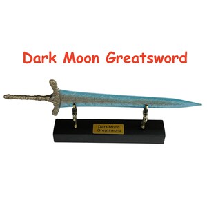 Op de afbeelding: Een decoratief groot zwaard met een blauw lemmet en een sierlijk handvat, tentoongesteld op een zwarte standaard. De tekst "Dark Moon Greatsword" is zichtbaar op het lemmet en het naamplaatje van de standaard.