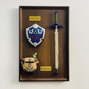 Può includere: Un'esposizione in legno incorniciata con uno Scudo Hylian, una Master Sword e una borsa marrone. Lo scudo è blu, bianco e rosso. La spada ha una lama argentata e un'impugnatura verde. La borsa è marrone con una pianta verde.