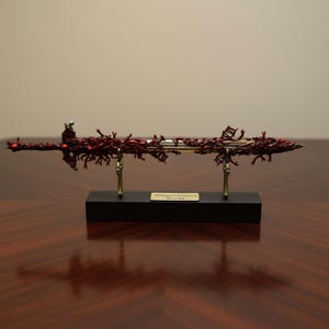 Può includere: Una spada dettagliata e ornata con un design rosso dall'aspetto organico. La lama è sostenuta da due supporti color oro su una base nera. Il testo "Blasphemous Blade" è su una piccola targa.