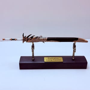 May include: A decorative long sword with a bronze blade and handle, displayed on a black wooden stand. The sword has intricate details and the text "Rathalos Long Sword" on a gold plaque.