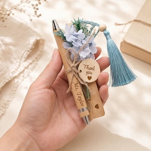 Può includere: Una penna a sfera in legno con dettagli in metallo argentato, incisa con il nome Ms. Taylor. La penna è decorata con fiori in tessuto azzurro, una nappa blu abbinata e una piccola etichetta rotonda in legno con il testo Thank you e un cuore.