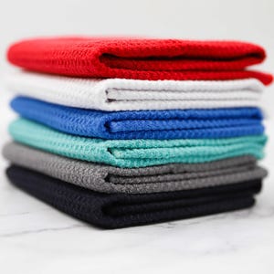 Puede incluir: Una pila de seis toallas de tejido de waffle en varios colores, incluyendo rojo, blanco, azul, verde azulado, gris y negro.