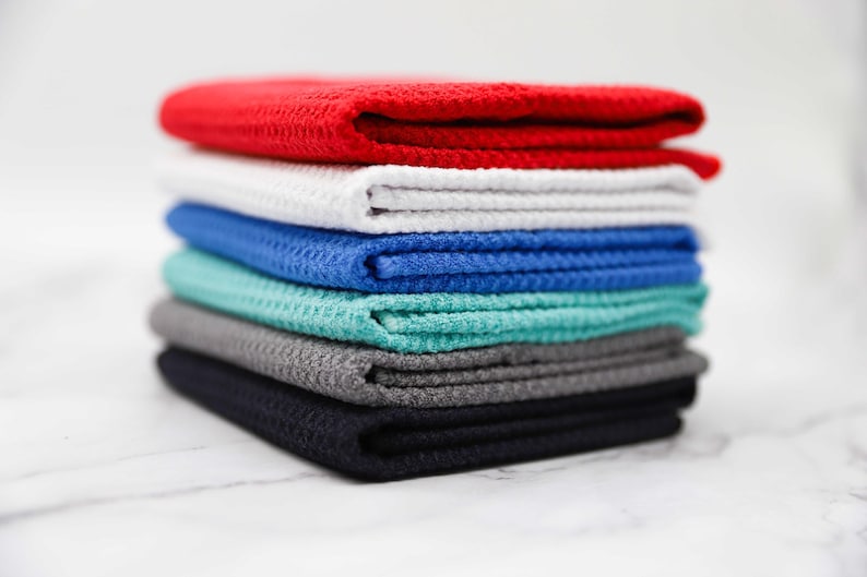 Puede incluir: Una pila de seis toallas de tejido de waffle en varios colores, incluyendo rojo, blanco, azul, verde azulado, gris y negro.