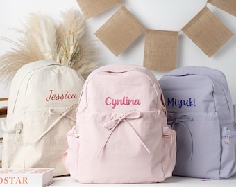 Op de afbeelding: Drie gepersonaliseerde rugzakken in crème, roze en lavendel. Elke rugzak heeft een geborduurde naam op de voorkant: "Jessica" in rood, "Cyntina" in roze en "Miyuki" in blauw. De rugzakken hebben een strikdetail en zijzakken.