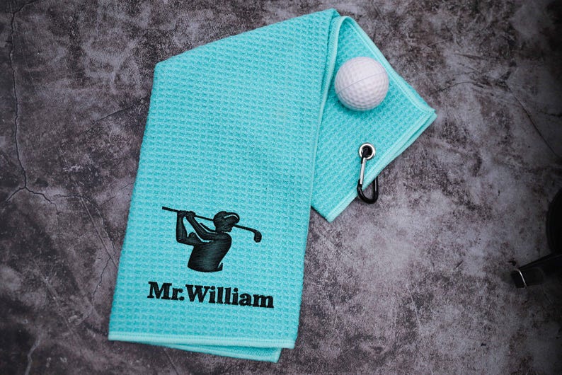 Puede incluir: Una toalla de golf azul claro con una silueta negra de un golfista y el texto "Mr. William" bordado en ella. Una bola de golf blanca est&aacute; sobre la toalla.