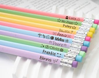 May include: A collection of colorful pencils with personalized names and small illustrations. Each pencil has a different color, including pink, green, yellow, and blue. The pencils are arranged in a row, with the erasers facing the same direction.