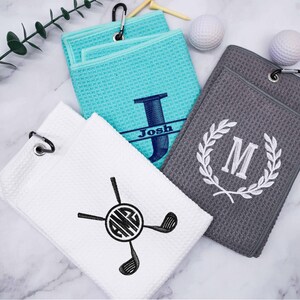 Puede incluir: Tres toallas de golf personalizadas con diferentes dise&ntilde;os. Una toalla es turquesa con la letra "J" y el nombre "Josh" bordado en ella. Otra toalla es gris con una corona blanca y la letra "M" bordada en ella. La tercera toalla es blanca con un palo de golf negro y monograma con las letras "AMG".