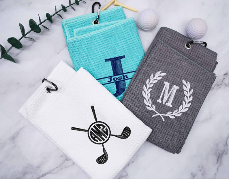 Puede incluir: Tres toallas de golf personalizadas con diferentes dise&ntilde;os. Una toalla es turquesa con la letra "J" y el nombre "Josh" bordado en ella. Otra toalla es gris con una corona blanca y la letra "M" bordada en ella. La tercera toalla es blanca con un palo de golf negro y monograma con las letras "AMG".