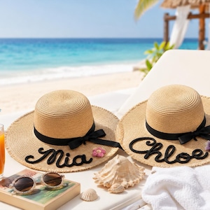 May include: Two tan straw sun hats with black ribbon bands and bows. One hat features the name Mia in black script, and the other features the name Chloe in black script. Each hat is decorated with a small purple crocheted flower on the brim.