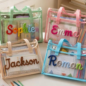 May include: Four rectangular clear plastic tote bags with fabric handles and snap closures. Each bag features a personalized name in raised, colorful yarn lettering on the front: Sophie, Emma, Jackson, and Roman. The bags contain notebooks and pencils.