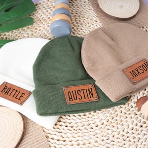 Puede incluir: Tres gorros de punto en blanco, verde oliva y beige, cada uno con un parche rectangular de cuero. Los parches están grabados con los nombres "BATTLE", "AUSTIN" y "JAXSON". Los sombreros están colocados sobre una estera tejida con juguetes de madera.