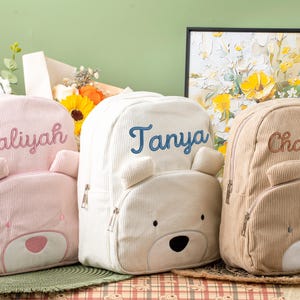 Może przedstawiać: Trzy sztruksowe plecaki w kolorze różowym, białym i beżowym, każdy z motywem misia i imieniem wyhaftowanym z przodu. Różowy plecak ma napis "Aaliyah", biały "Tanya", a beżowy "Chantal".