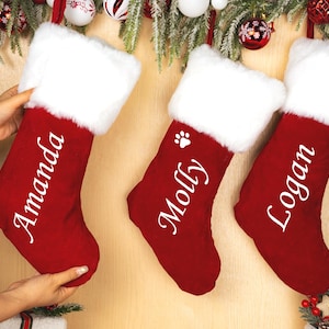 Könnte beinhalten: Drei rote Weihnachtsstrümpfe mit weißem Kunstfellbesatz und personalisierten Namen: Amanda, Molly und Logan. Der Molly-Strumpf hat einen Pfotenabdruck. Festliche Weihnachtsdekoration.