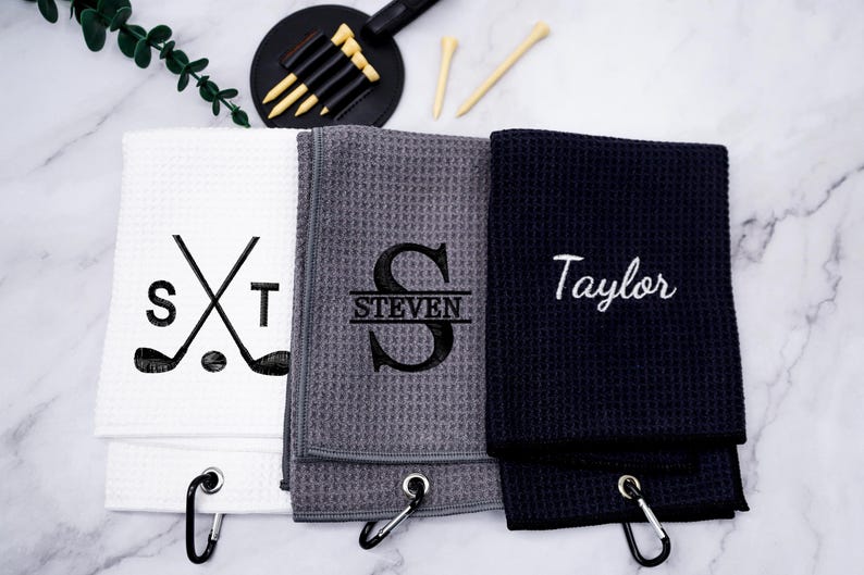 Puede incluir: Tres toallas de golf con bordado personalizado. La primera toalla es blanca con bordado negro de palos de golf cruzados y las iniciales "ST". La segunda toalla es gris con bordado negro de la letra "S" y el nombre "STEVEN" debajo. La tercera toalla es negra con bordado blanco del nombre "Taylor".