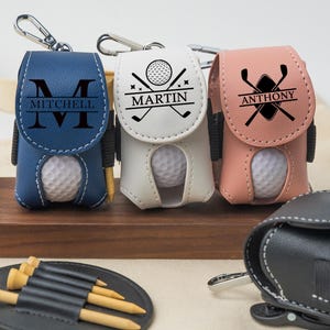 Op de afbeelding: Drie gepersonaliseerde golfbalhouders met elk een golfbal erin. De houders zijn gemaakt van leer en hebben verschillende kleuren en ontwerpen. De eerste houder is blauw met de naam "Mitchell" erop. De tweede houder is wit met de naam "Martin" en een golfclubontwerp. De derde houder is roze met de naam "Anthony" en een golfclubontwerp.