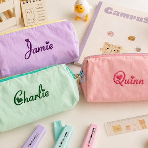 Op de afbeelding: Drie rechthoekige etuis van stof in de kleuren lavendel, mintgroen en lichtroze. Elk etui is voorzien van een ritssluiting en een geborduurde naam: het lavendelkleurige etui toont Jamie, het mintgroene Charlie en het roze etui Quinn.