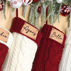 May include: Four Christmas stockings in red and white cable knit with personalized leather name tags. The names Sophia, Agnes, Bella, and Justin are displayed. The stockings are decorated with festive ornaments and greenery.