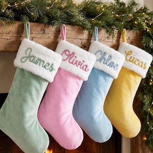 May include: Four velvet Christmas stockings in pastel green, pink, blue, and yellow. Each stocking features a white faux fur cuff embroidered with the names James, Olivia, Chloe, and Lucas in matching thread colours, hanging from a wooden mantel.