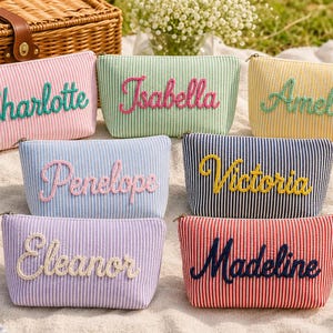 May include: Six rectangular zipped pouches made of striped fabric in pink, light green, yellow, light blue, dark blue, and red. Each pouch features a name embroidered in a cursive font: Charlotte, Isabella, Amelia, Penelope, Victoria, Eleanor, and Madeline.