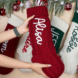 Puede incluir: Calcetines navideños de punto trenzado rojos, blancos y verdes, cada uno con un nombre bordado en blanco. Los calcetines tienen un puño en la parte superior. Nombres: Alisa, Claire, JFC y Rolie. Decoración festiva navideña.