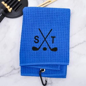 Puede incluir: Toalla de golf azul con un dise&ntilde;o bordado en negro de palos de golf cruzados y las letras "S" y "T".