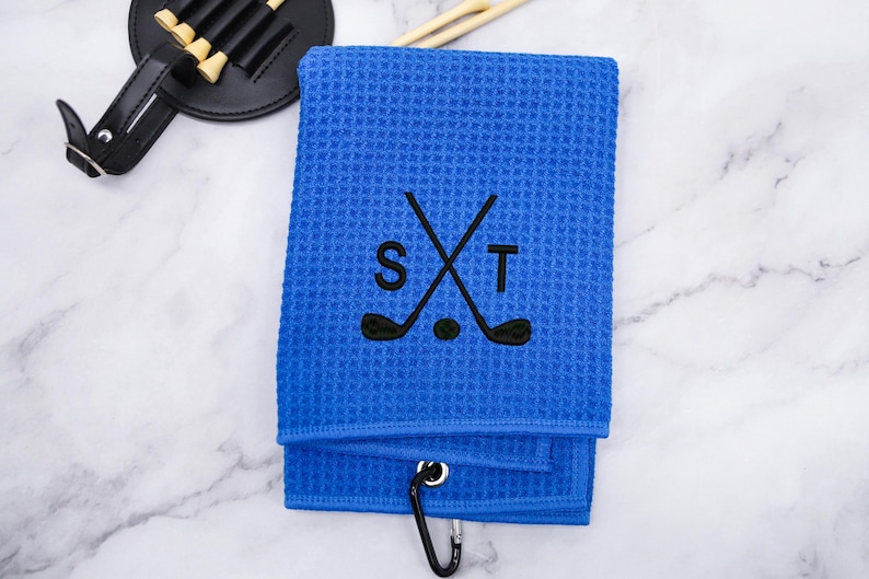 Puede incluir: Toalla de golf azul con un dise&ntilde;o bordado en negro de palos de golf cruzados y las letras "S" y "T".