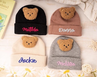 May include: Four knitted beanies in black, mauve, cream, and gray, each with a teddy bear face applique. The names "Matilda", "Donna", "Jackie", and "Melissa" are embroidered on the front in pink and blue.