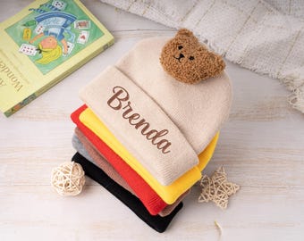 May include: A stack of colorful knit beanies, each folded neatly. The top beanie is beige with the name "Brenda" embroidered in brown. A small brown teddy bear head is attached. Other beanies are red, yellow, gray, brown, and black.