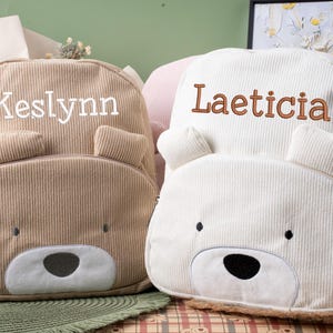 May include: Two corduroy backpacks in the shape of bears. One is tan with the name "Keslynn" embroidered in white, and the other is off-white with "Laeticia" embroidered in brown. Both have bear faces.