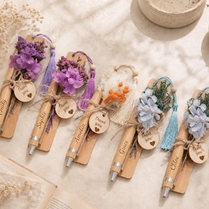 May include: Five wooden pens attached to rectangular wooden bookmarks, each personalised with a name and decorated with dried flowers, a fabric tassel, and a small round wooden tag engraved with the words Thank you and a heart symbol.